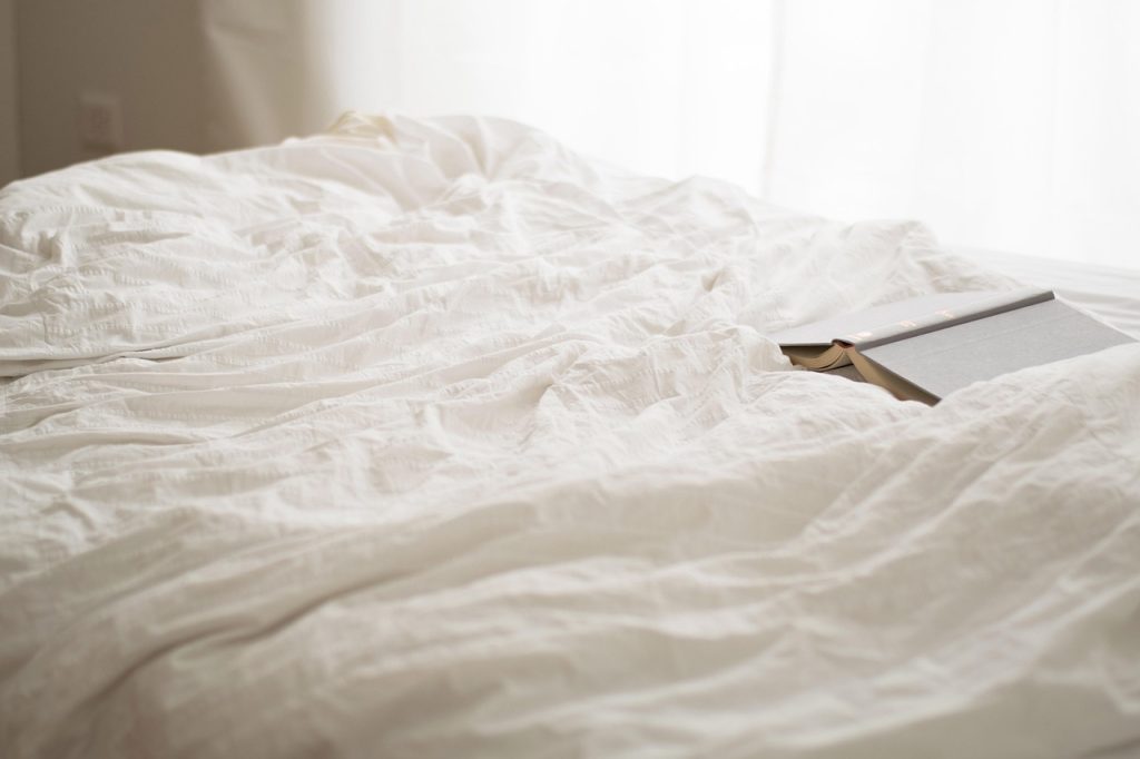 An open book on rumpled white bedding beside a sunlit window.
