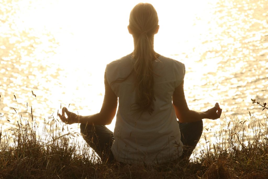 A woman sitting cross-legged in a meditation pose by a sunlit lake at golden hour.