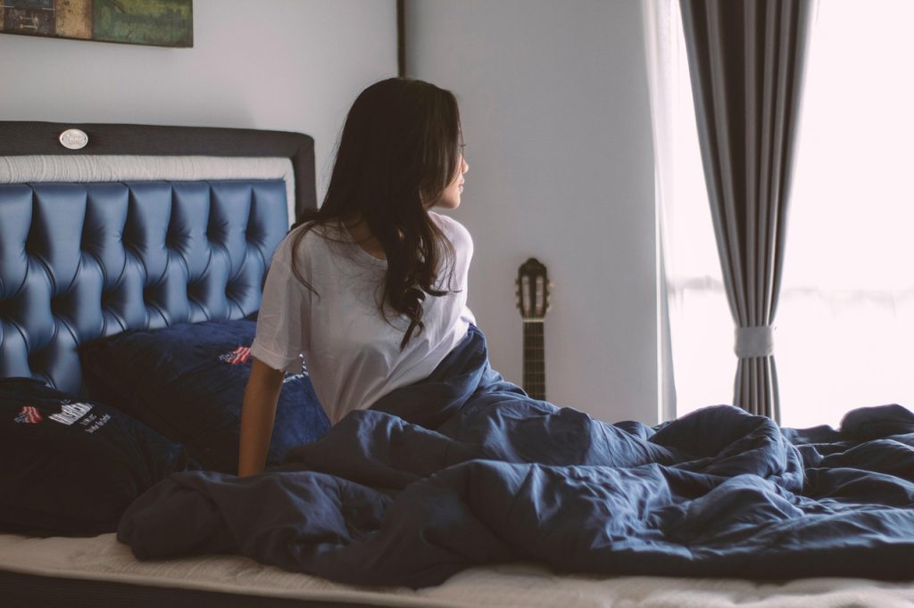 A woman with long dark hair sitting on a bed and looking toward a bright window.