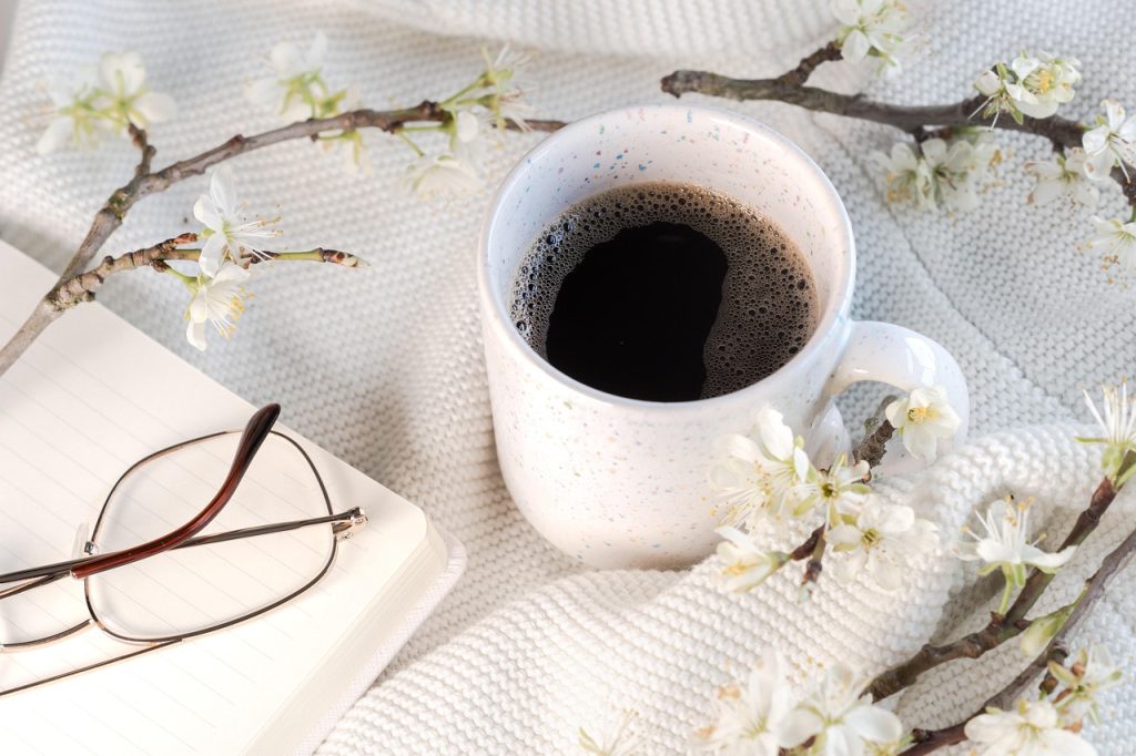 A cup of black coffee, an open notebook with reading glasses, and white blossom branches on a knit blanket.