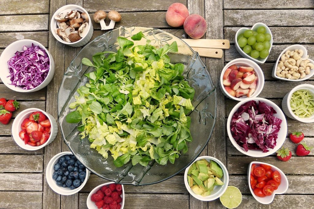 A bowl of green salad surrounded by small bowls of chopped vegetables, fruit, berries and nuts on a wooden table.
