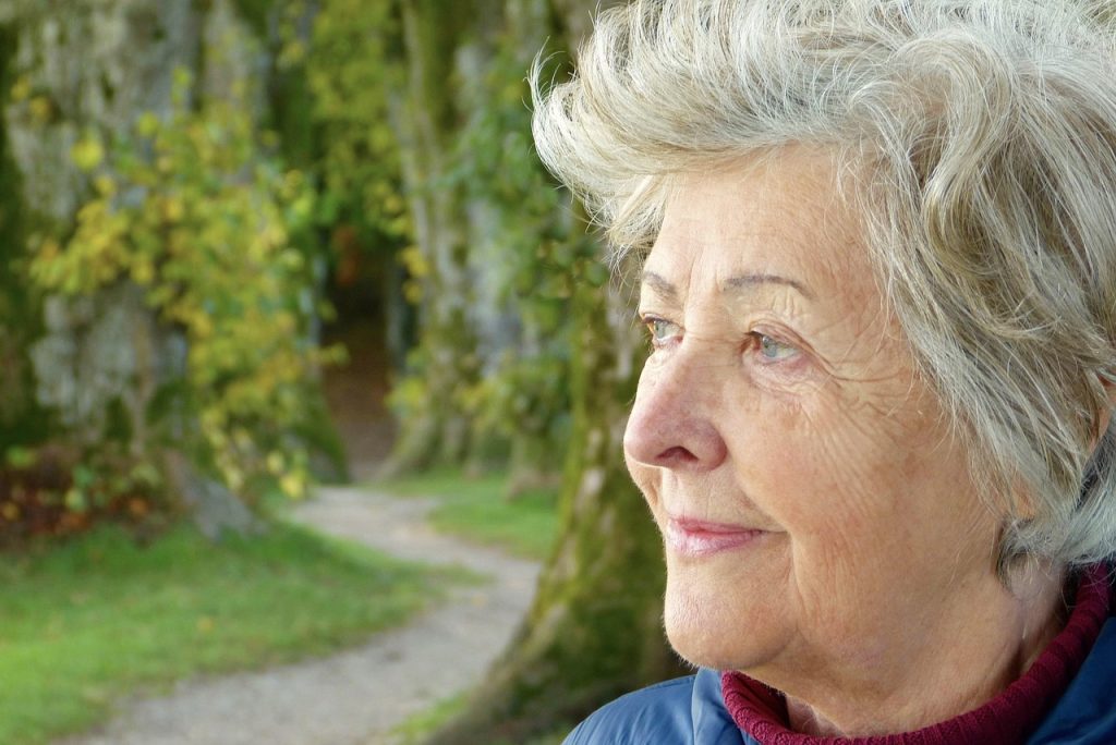 An older woman with grey hair looking into the distance along a wooded path.