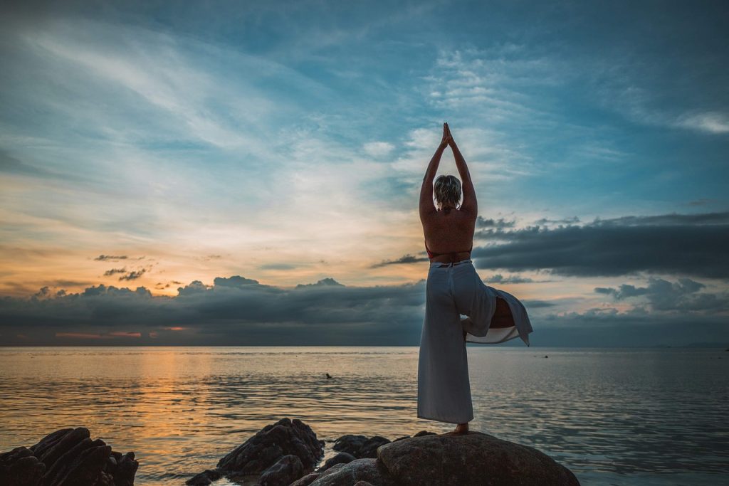 A woman in a yoga pose on a rock beside the sea at dusk.