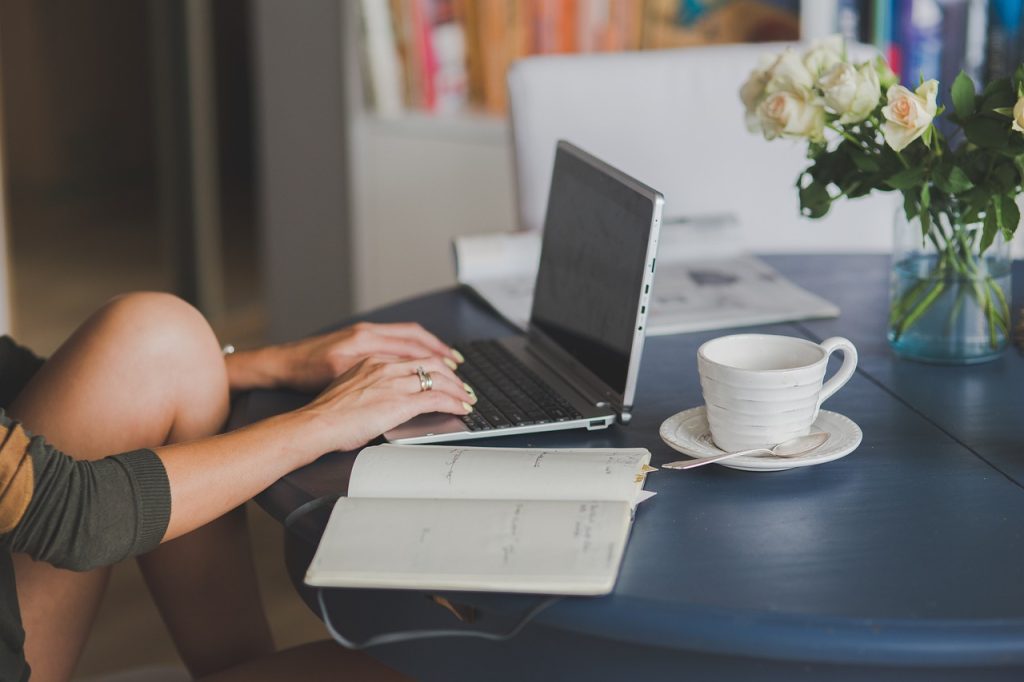 A woman typing on a laptop at a table with an open notebook, a cup of coffee and a vase of roses.