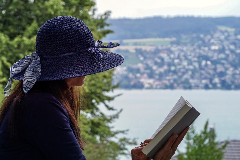 A woman in a sun hat reading a book beside a lake.