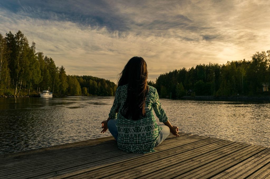 A woman sitting cross-legged on a wooden dock overlooking a lake at sunset.