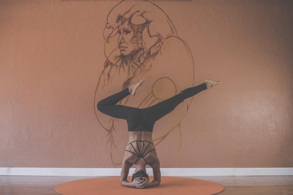 A woman in a headstand yoga pose on a mat in front of a wall with a large pencil sketch.