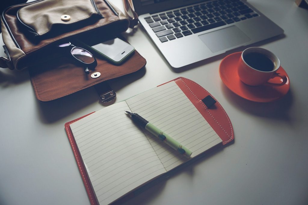 An open notebook with a pen beside a laptop, coffee cup, glasses and a bag on a desk.