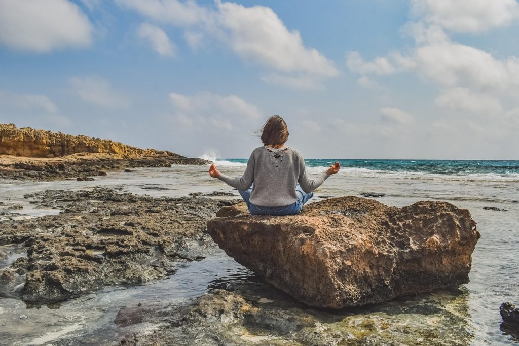 A woman sitting cross-legged on a rock by the sea, seen from behind.