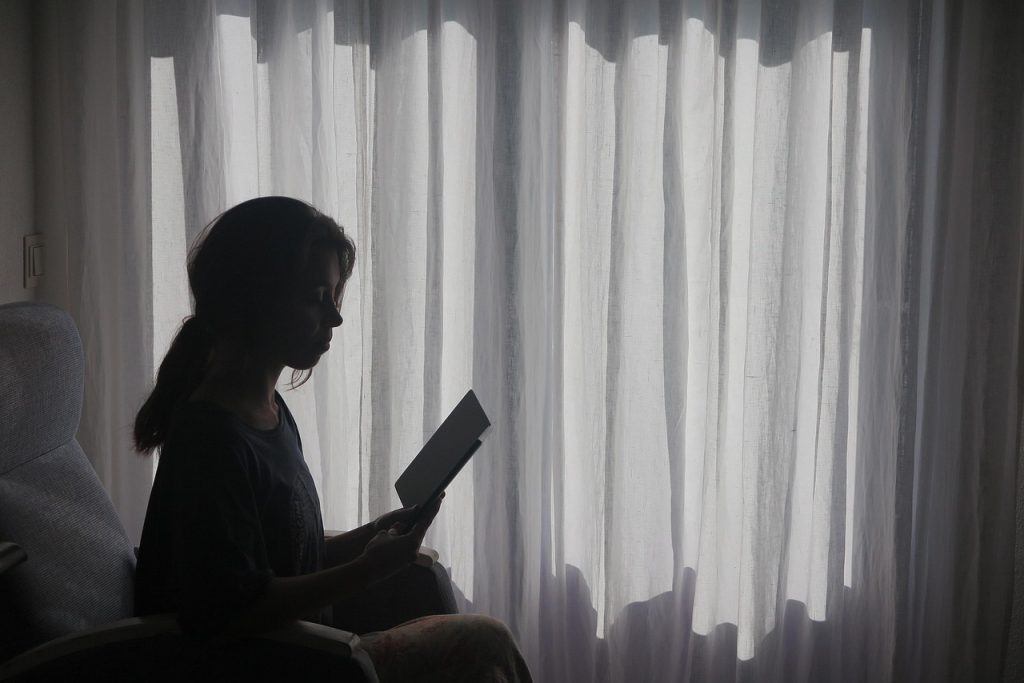 Silhouette of a woman sitting and reading beside a sheer-curtained window.