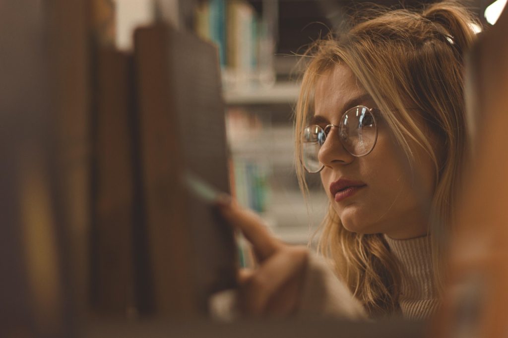 Woman wearing round glasses browsing books on a library shelf.