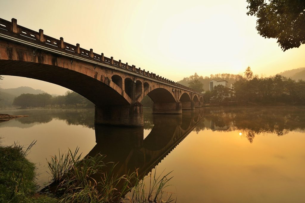 Stone arch bridge reflected in calm water at sunrise.
