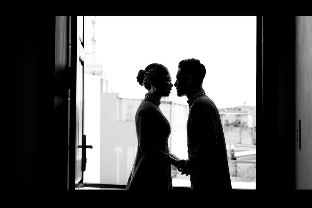 Silhouette of two adults standing face to face and holding hands by a bright window.