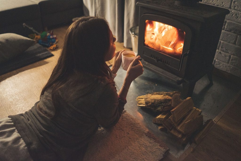 Woman resting on the floor holding a mug beside a wood-burning stove.