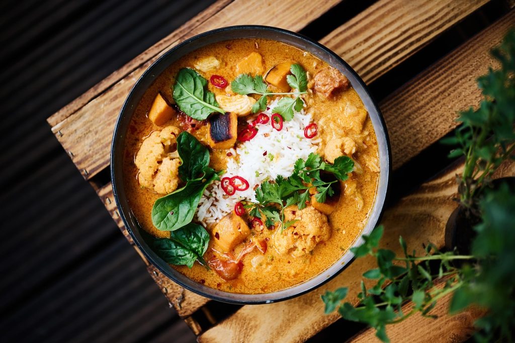Bowl of curry with rice, vegetables and fresh herbs on a wooden surface.