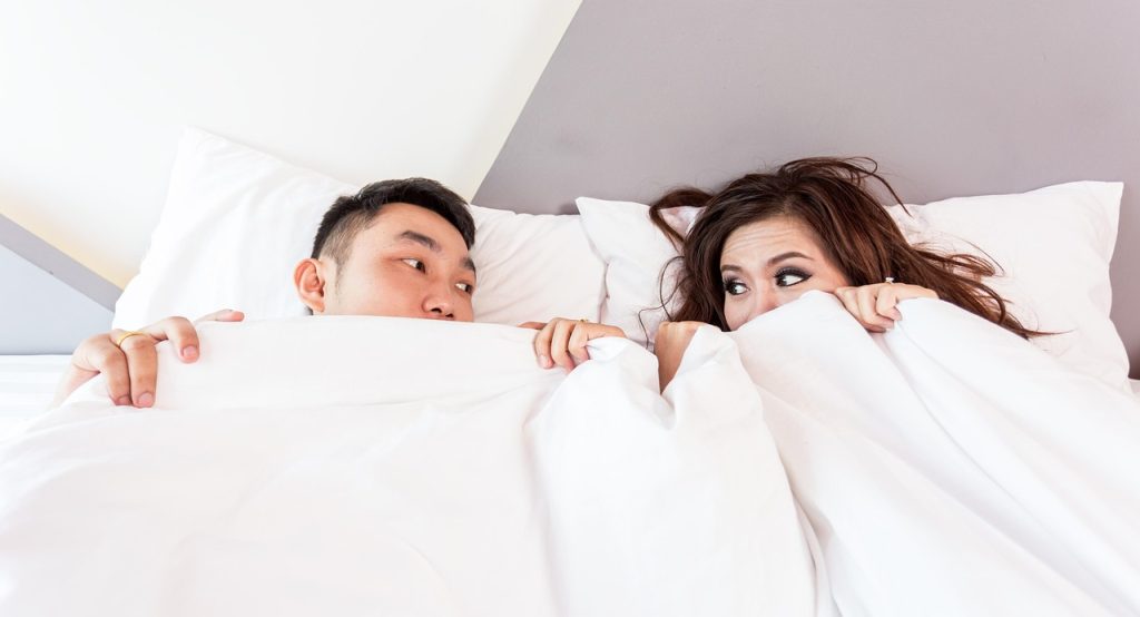 Two adults lying in bed peeking over a white duvet.