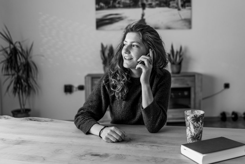 Black-and-white photo of a woman sitting at a table talking on a phone, with a cup and notebook nearby.