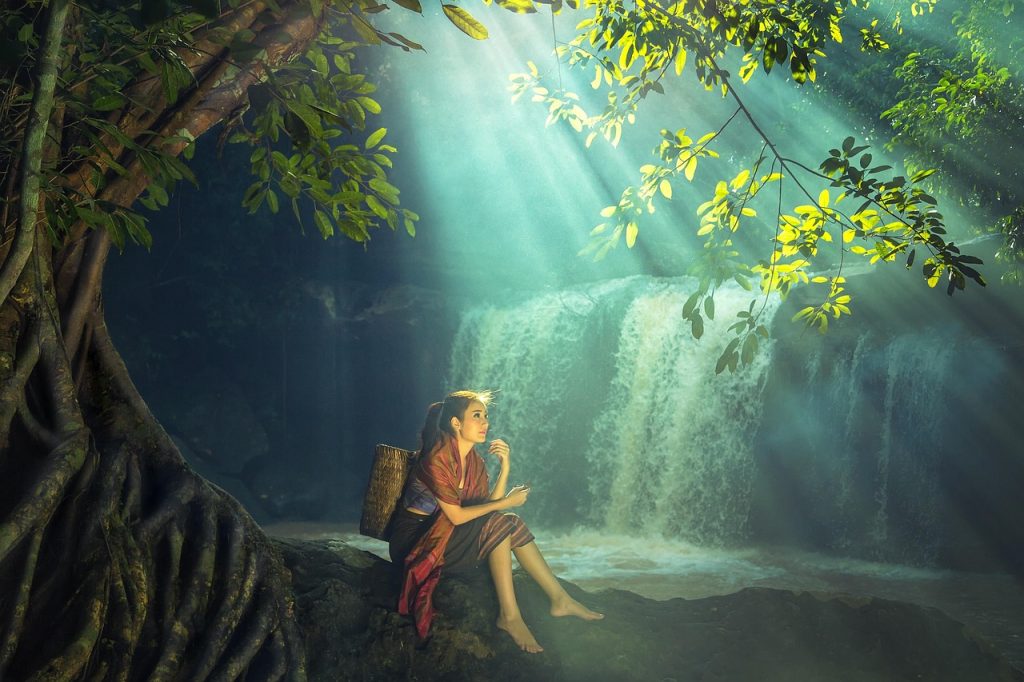 Woman sitting on a rock beside a forest waterfall as sunbeams filter through the trees.