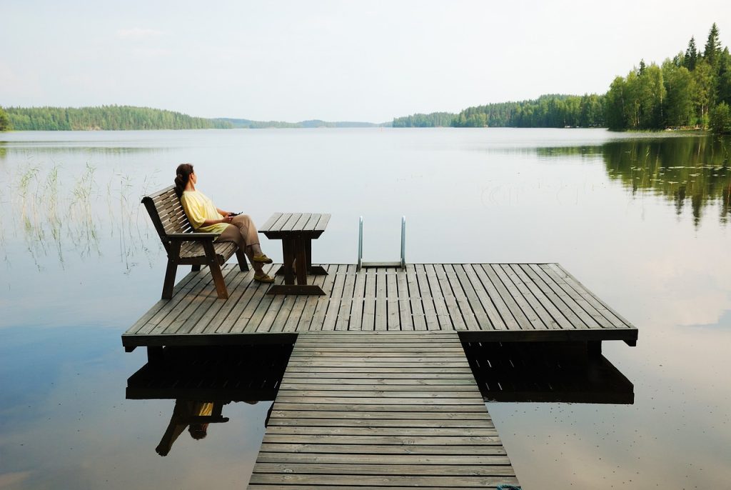 Person sitting on a bench on a wooden dock beside a calm lake surrounded by forest.