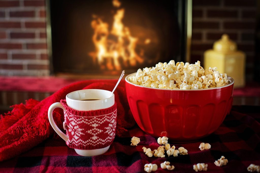 Bowl of popcorn and a mug of tea on a blanket in front of a lit fireplace.
