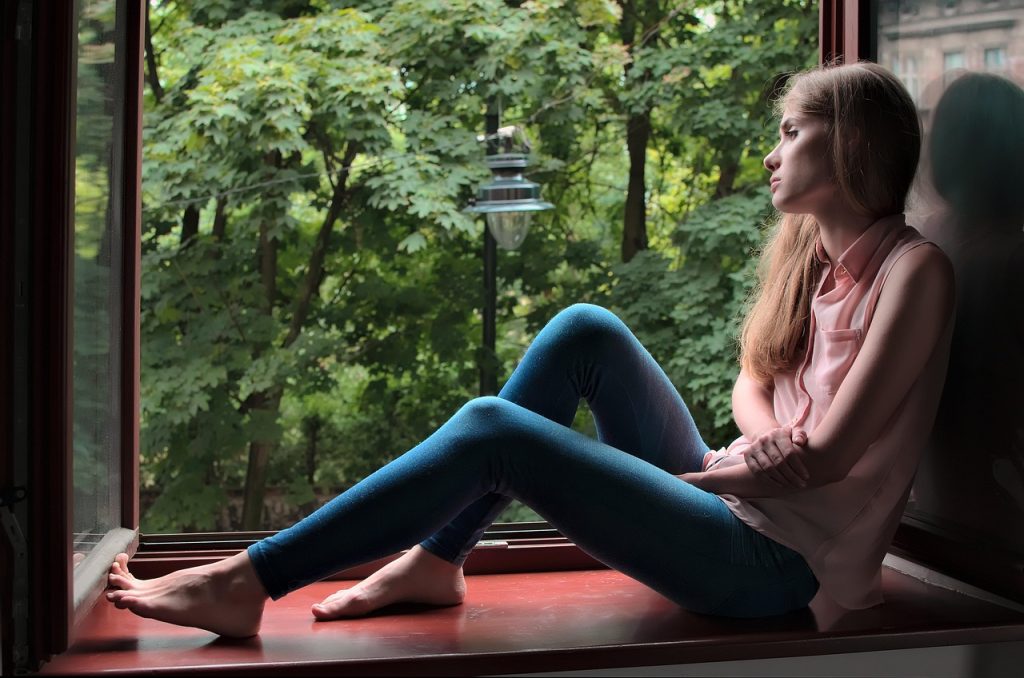 Woman sitting barefoot on a windowsill looking out at green trees.