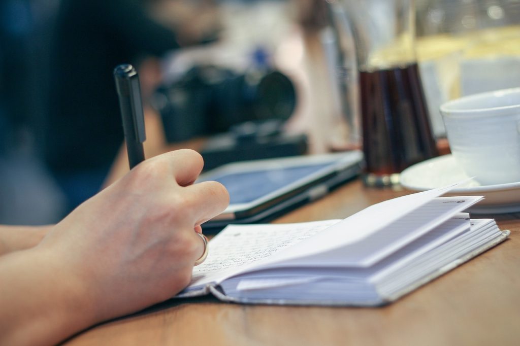 A hand writing in a small notebook at a wooden table, with a cup and camera blurred behind.