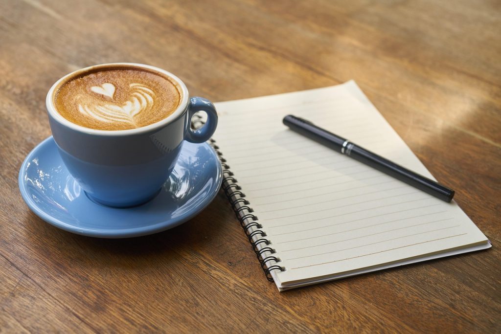 A cup of coffee with latte art beside an open spiral notebook and a pen on a wooden table.