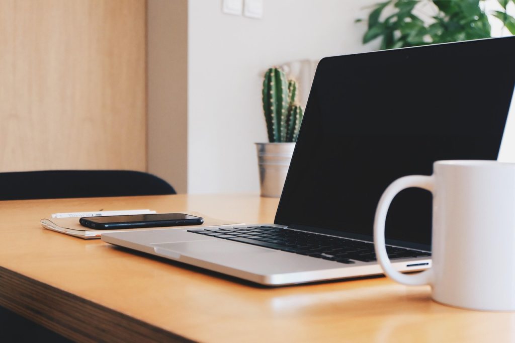 An open laptop, smartphone and notebook on a wooden desk beside a white mug and a small potted cactus.
