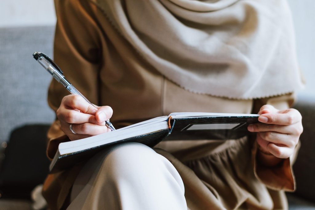 A woman in a beige headscarf writing in a notebook with a pen while seated.