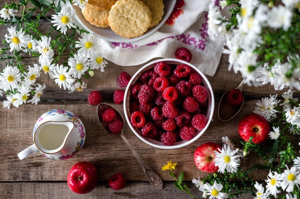 A bowl of fresh raspberries on a rustic wooden table surrounded by daisies, apples and a small jug of cream.