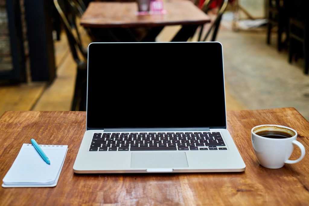 A laptop with a blank screen beside a small notepad and a cup of coffee on a wooden table.