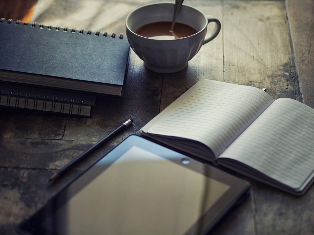 An open blank notebook and a tablet beside a cup of tea on a wooden table.