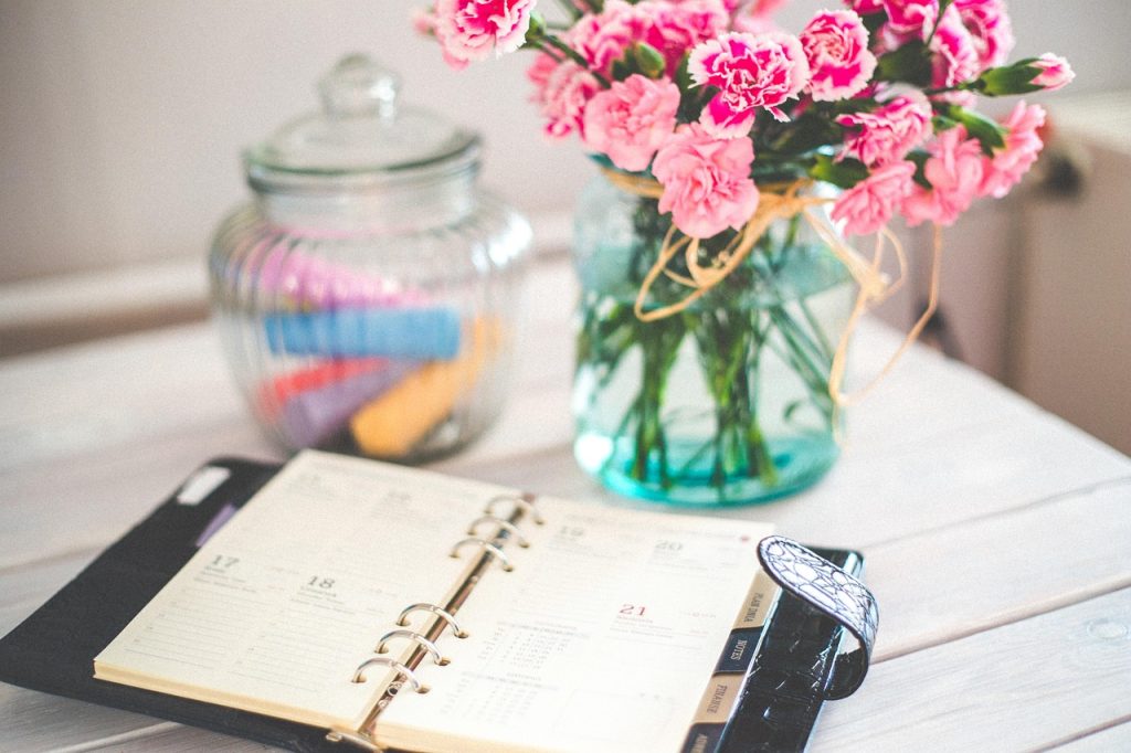 An open ring-bound planner beside a jar of pink carnations on a white table.