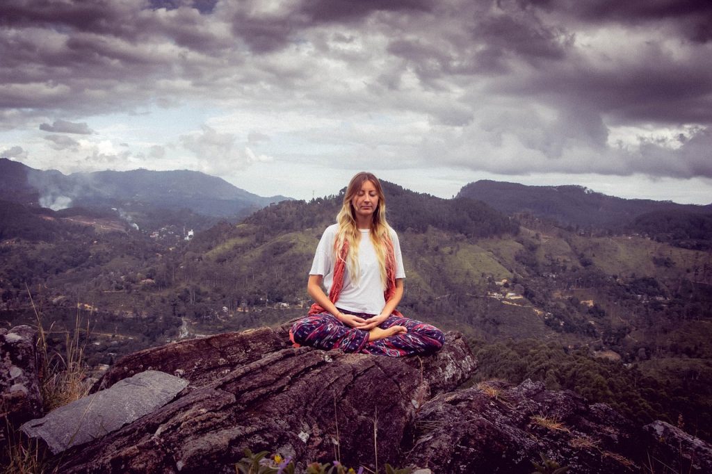 A woman sitting cross-legged in a meditation pose on a rock, with hills in the background.
