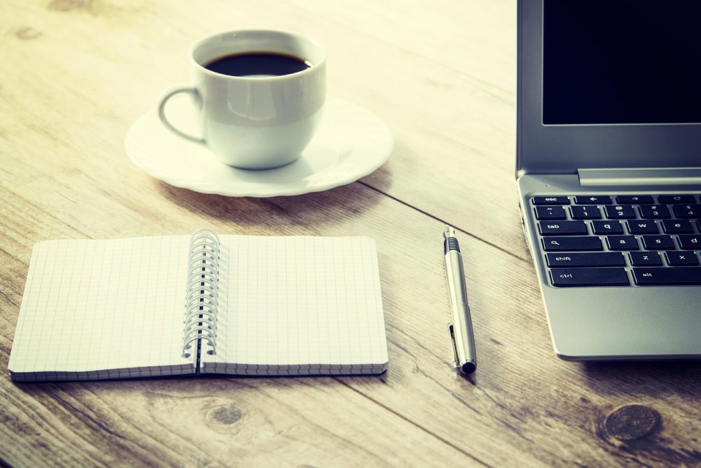 An open blank notebook and a pen beside a cup of coffee and a laptop on a wooden table.