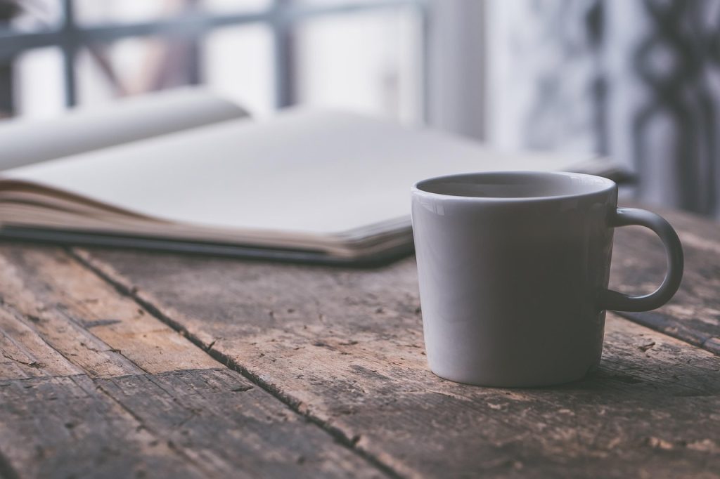 A white mug on a rustic wooden table beside an open blank notebook near a window.