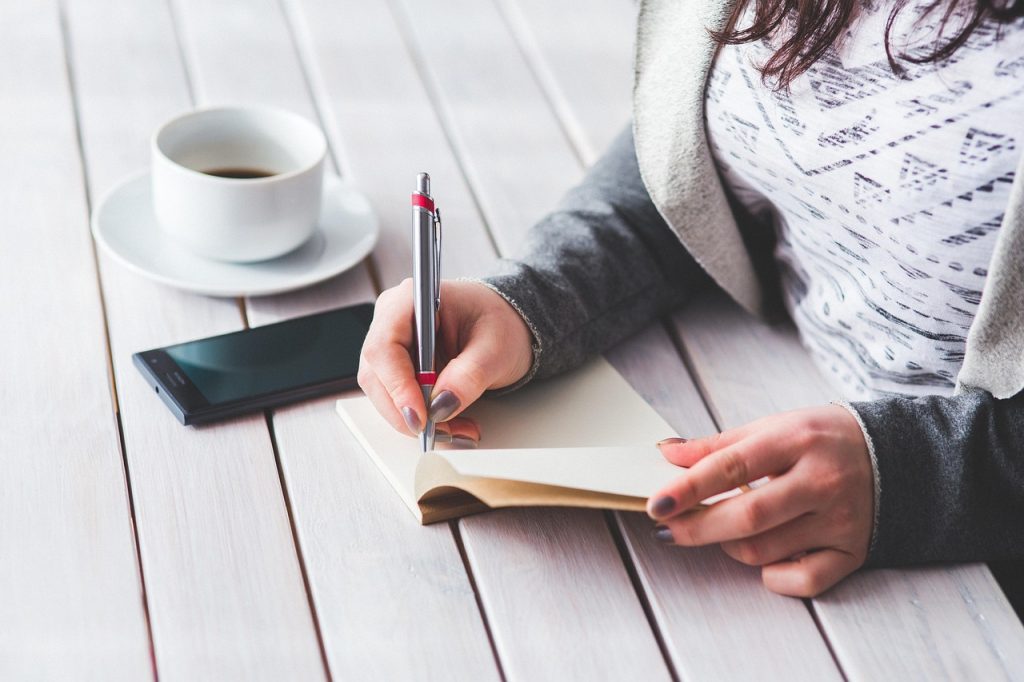 A woman writing in a notebook beside a cup of coffee and a phone on a table.