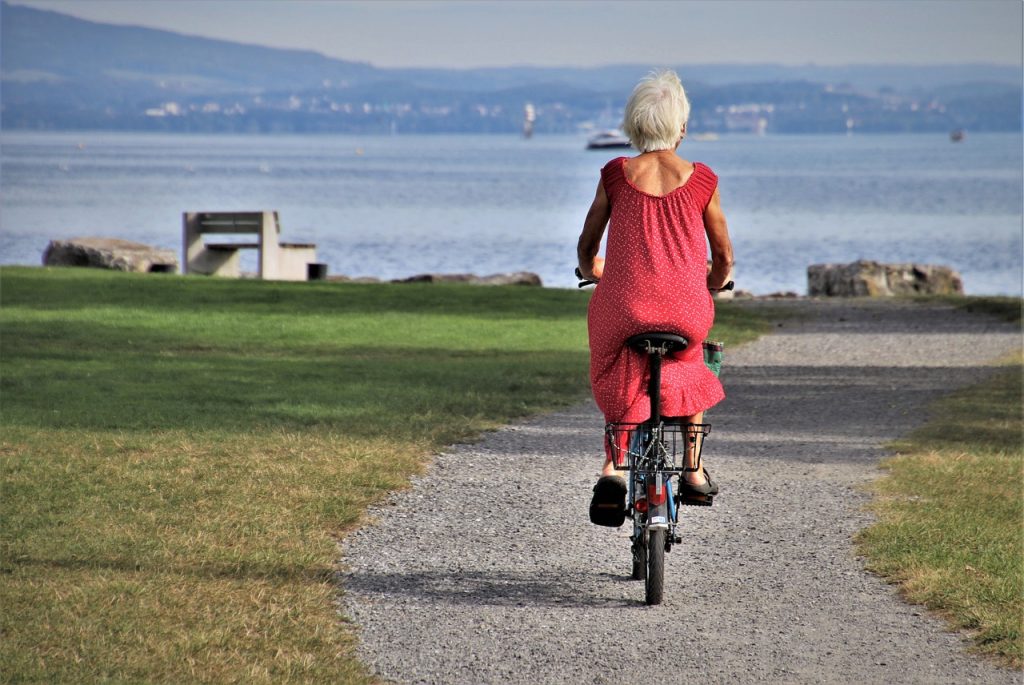 An older woman in a red dress riding a bicycle along a waterfront path.
